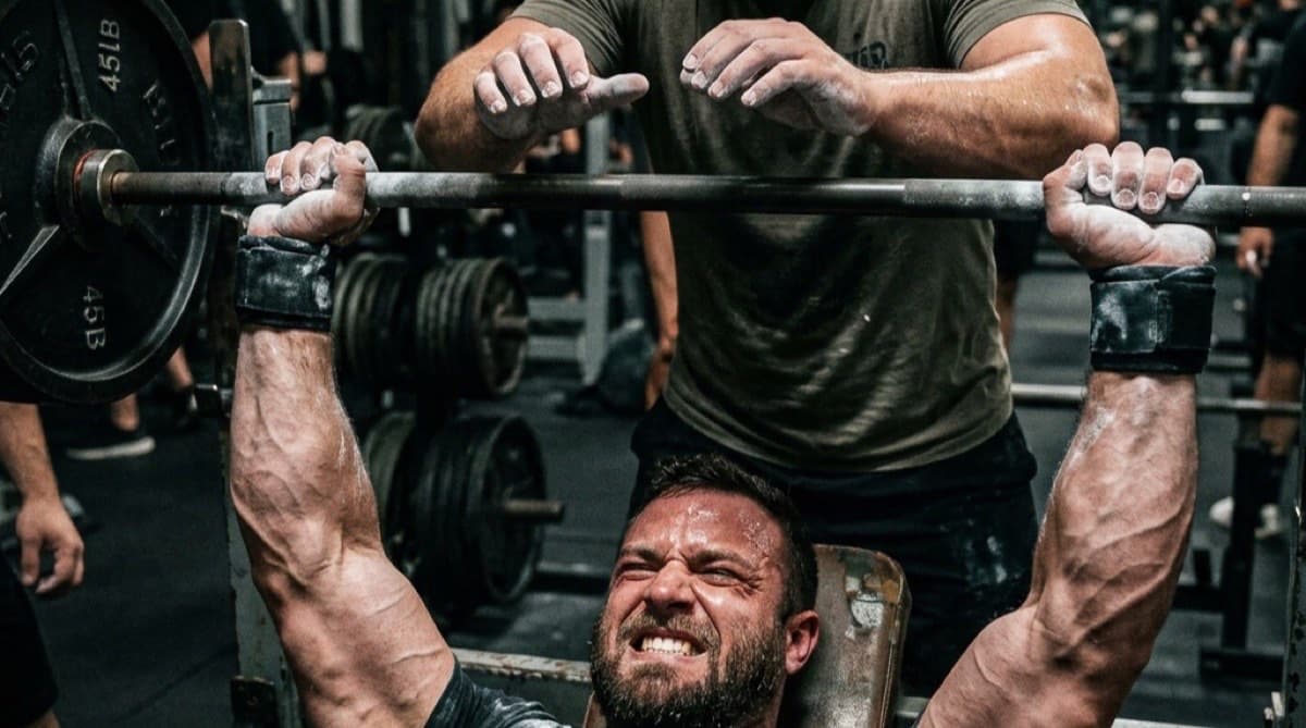 A lifter straining through the final rep of a barbell bench press with a spotter hovering hands above the bar in a dimly lit gym.